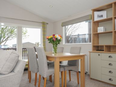 A dining area with a wooden table and four gray chairs with a vase of flowers on the table and a wooden cabinet in the corner at Lodge 67 at Riviera Bay Coastal Retreat Brixham