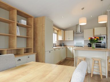 A kitchen with wooden shelves cabinets a countertop with stools and flowers and a dining table with chairs at Lodge 67 at Riviera Bay Coastal Retreat in Brixham