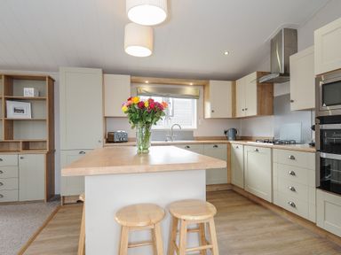 A kitchen with an island, wooden stools, cabinets, a stove, microwave, and a vase of flowers on the island at Lodge 67 at Riviera Bay Coastal Retreat in Brixham