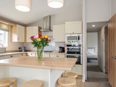 A kitchen with a wooden island table and stools a vase of flowers built in oven and a view into a bedroom at Lodge 67 at Riviera Bay Coastal Retreat in Brixham