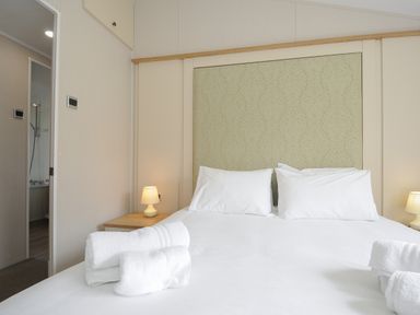 A bedroom with a bed two pillows bedside tables with lamps and rolled towels near an open door to a bathroom at Lodge 67 at Riviera Bay Coastal Retreat in Brixham