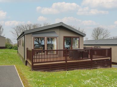 A single story cabin with brown wooden deck surrounded by grass and trees at Lodge 67 at Riviera Bay Coastal Retreat in Brixham