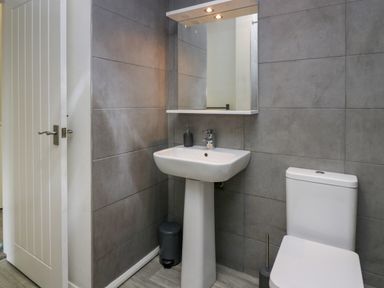 A bathroom with a white pedestal sink and toilet grey tiled walls and a mirror cabinet with lights above at Acorn Cottage in Windermere