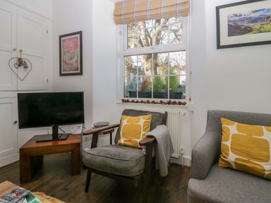 A living room with a grey armchair and sofa with yellow cushions a wooden side table with a TV and a window with pinecones on the sill at Acorn Cottage in Windermere