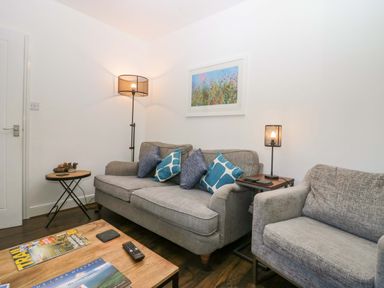 A living room with a gray sofa and armchair with blue cushions a floor lamp and small tables with lamps and magazines at Acorn Cottage in Windermere