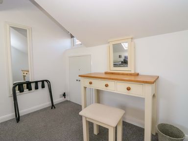 A small room with a wooden dressing table with mirror and stool a tall wall mirror a black luggage rack and a basket at Acorn Cottage in Windermere