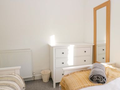 A bedroom corner with a white chest of drawers next to a tall mirror and a bed with a blanket and towel at Acorn Cottage in Windermere