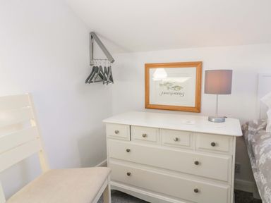 A bedroom corner with a white chair a white dresser with a lamp and framed picture on the wall and hangers on a wall-mounted rack at Acorn Cottage in Windermere