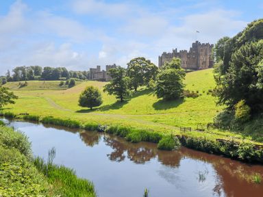 A river with grass and trees beside a castle on a hill at The Wizard's Chamber in Alnwick
