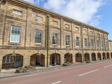 A stone building with arched doorways and large windows along a street with flower pots hanging and two people sitting on a bench at The Wizard's Chamber in Alnwick