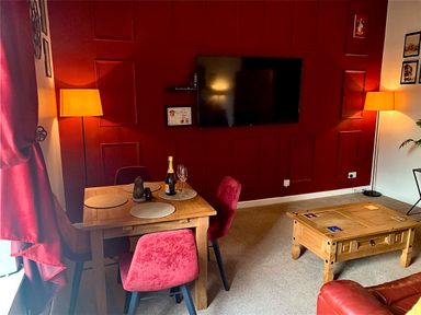 A dining area with a wooden table and four red chairs next to a wall with a mounted TV and two floor lamps at The Wizard's Chamber in Alnwick