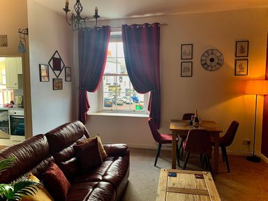 A living room with a leather sofa red curtains a dining table with four chairs and a floor lamp at The Wizard's Chamber in Alnwick