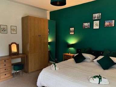 A bedroom with a bed pillows bedside table lamp wardrobe dressing table and framed pictures on a green wall at The Wizard's Chamber in Alnwick