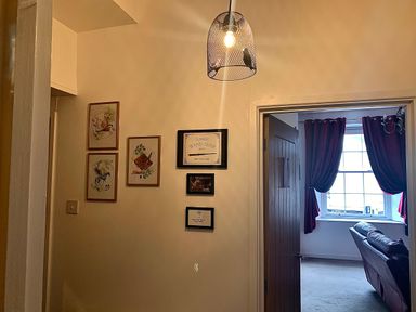 A hallway with framed pictures on the wall and a pendant light fixture next to a room with a window red curtains and a couch at The Wizard's Chamber in Alnwick