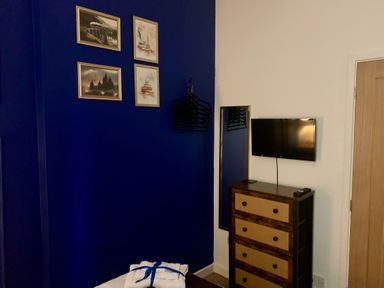 A room corner with a dresser, wall-mounted TV, mirror, paintings on a blue wall, and hangers at The Wizard's Chamber in Alnwick