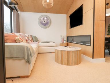 A living room with a white sectional sofa colorful cushions a round wooden coffee table a wall-mounted TV and a modern fireplace at Encil Gyfrinfa in Gwynus near Llithfaen