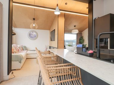 An open kitchen and living area with a countertop, rattan bar stools, a sofa, a wall clock, and a television at Encil Gyfrinfa in Gwynus near Llithfaen