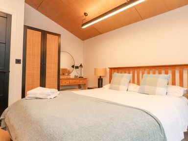 A bedroom with a wooden bed and headboard two large checked pillows a grey blanket a wooden dressing table with a round mirror and a lamp next to a wardrobe at Encil Gyfrinfa in Gwynus near Llithfaen