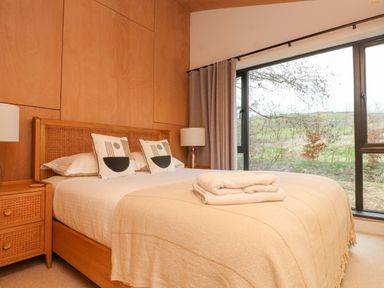 A bedroom with a wooden bed frame bedside tables lamps and large windows showing an outdoor view at Encil Gyfrinfa in Gwynus near Llithfaen