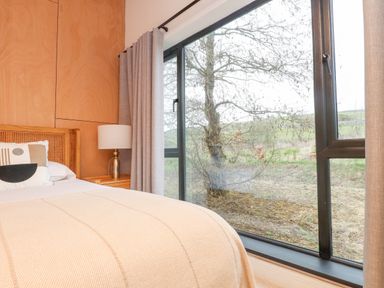A bedroom with a bed a bedside table with a lamp and a large window showing an outdoor tree at Encil Gyfrinfa in Gwynus near Llithfaen