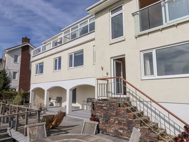 An outdoor decking area with wooden tables and chairs in front of a multi-story building with arches and multiple windows at Seahaze in Downderry