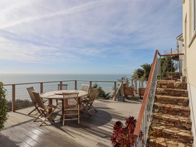 An outdoor wooden deck with a round table and six chairs overlooking the sea at Seahaze in Downderry