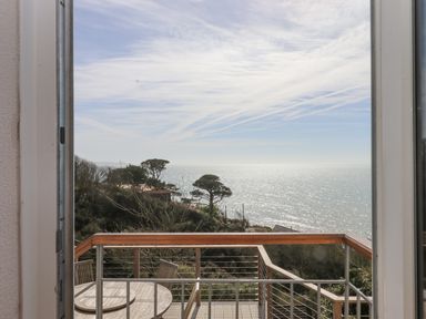 A balcony with wooden table and chairs overlooking trees and the ocean at Seahaze in Downderry