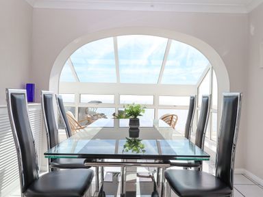 A dining room with a glass table and six black chairs near large windows with a view at Seahaze in Downderry