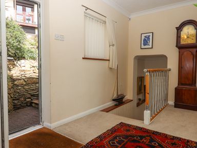 An indoor area with a staircase, a door leading outside, a tall wooden clock, a framed picture, and a patterned rug at Seahaze in Downderry