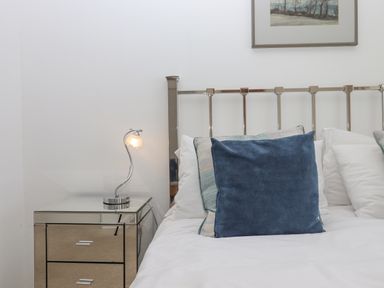 A bed with white sheets and blue and patterned pillows next to a mirrored nightstand with a modern lamp and a framed picture on the wall at Seahaze in Downderry