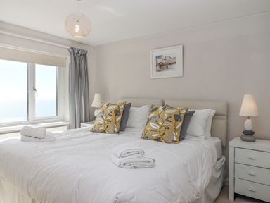 A bedroom with a double bed with white bedding and yellow patterned cushions two bedside tables with lamps a window with curtains and a framed picture on the wall at Seahaze in Downderry