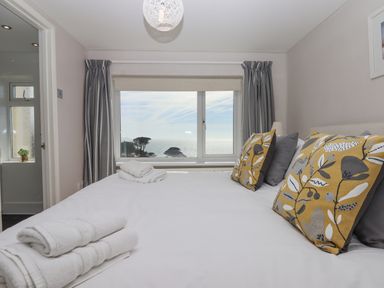A bedroom with a large bed with white linens and yellow patterned pillows overlooking the sea through a window at Seahaze in Downderry