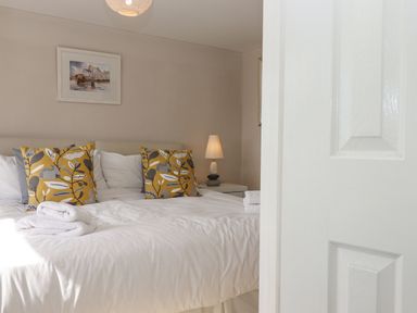 A bedroom with a bed with white bedding yellow patterned pillows folded towels on the bed a bedside table with a lamp and a painting on the wall at Seahaze in Downderry