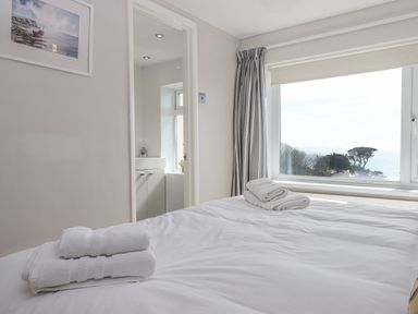 A bedroom with a bed covered in white linens and towels by a window with a view of trees and the sea at Seahaze in Downderry