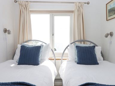A bedroom with two single beds with blue pillows next to a window with beige curtains at Seahaze in Downderry