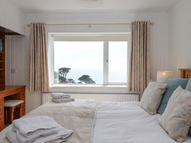 A bedroom with a bed, pillows, folded towels, a window with curtains, a lamp, and a wooden desk at Seahaze in Downderry