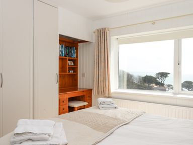 A bedroom with a bed and folded towels next to a wooden desk with shelves and a large window showing trees and water at Seahaze in Downderry