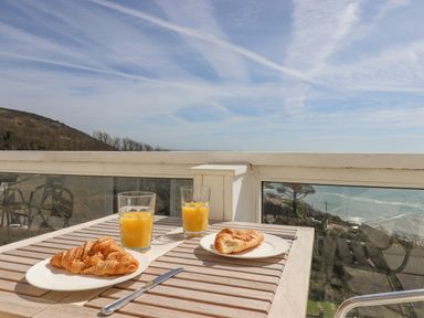 A wooden table with croissants and glasses of orange juice on a balcony overlooking hills and the sea at Seahaze in Downderry
