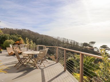 An outdoor wooden deck with tables and chairs overlooking trees and the ocean at Seahaze in Downderry
