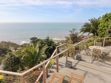 A wooden deck with chairs and a table overlooking the sea with trees and plants at Seahaze in Downderry