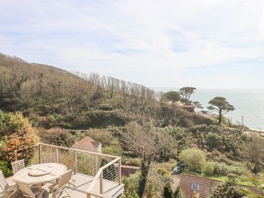 An outdoor deck with a round wooden table and chairs overlooking hillside vegetation and the sea at Seahaze in Downderry