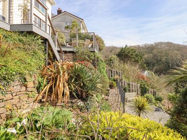 A garden with a paved path leading past plants and a wooden bench next to a house with a raised deck at Seahaze in Downderry