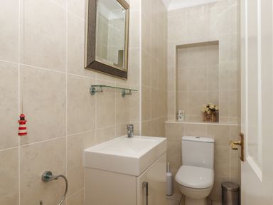 A bathroom with a toilet a sink a wall-mounted mirror and a small shelf with flowers at Seahaze in Downderry