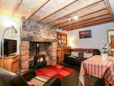 A living room with a stone fireplace black wood stove two black leather chairs a red checkered rug and a table with a checkered tablecloth at White Lilac Cottage in Brynsiencyn