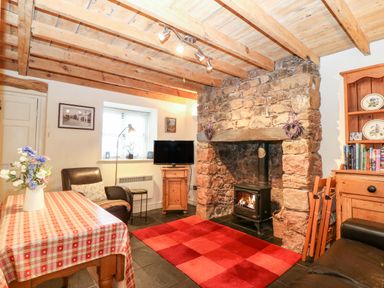 A living room with a stone fireplace wood burner red checkered rug table with flowers and wooden furniture at White Lilac Cottage in Brynsiencyn