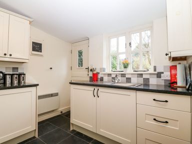 A kitchen with cream cabinets black countertops a window above the sink and a door at White Lilac Cottage Brynsiencyn