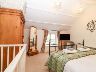 A bedroom with a metal bed frame towels on bed a wooden wardrobe a standing mirror a chair a television and a window with curtains at White Lilac Cottage in Brynsiencyn