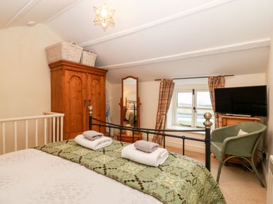 A bedroom with a double bed folded towels a wooden wardrobe a mirror a window with curtains a television and a green chair at White Lilac Cottage in Brynsiencyn
