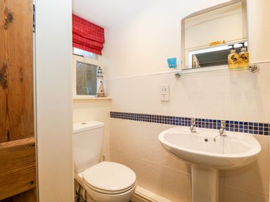A bathroom with a toilet a pedestal sink a mirror and a red window curtain at White Lilac Cottage in Brynsiencyn