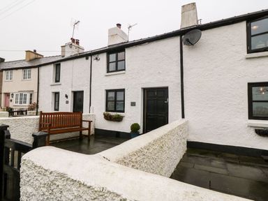 Exterior view of white stone terraced houses with black doors and windows a wooden bench and a small front patio at White Lilac Cottage in Brynsiencyn